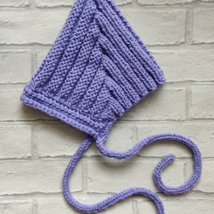 May include: A lavender knitted baby bonnet with a long tie. The bonnet is made with a simple stitch pattern and has a pointed crown.