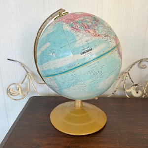 Może przedstawiać: Zabytkowy globus z jasnoniebieskim oceanem i kolorowymi lądami. Globus jest oznaczony jako "World Scholar Series" i stoi na złotej podstawie. Globus jest podtrzymywany przez metalową ramę.