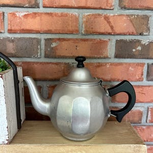 May include: A silver-colored teapot with a black handle and lid knob. The teapot has a rounded body and a curved spout. It sits on a wooden shelf in front of a brick wall.