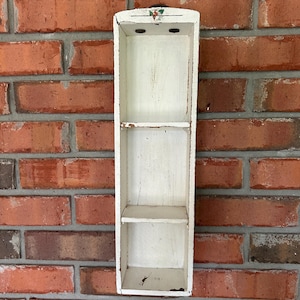May include: A tall, rectangular, white-painted wooden wall shelf featuring two internal horizontal shelves. The wood shows signs of wear with visible paint chipping. A small, painted holly leaf and berry motif is centered at the top edge.