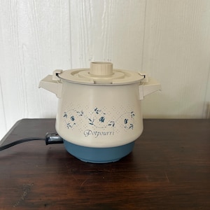 Op de afbeelding: Een elektrische potpourri-warmer met een crèmekleurige behuizing en een blauwe voet. De buitenkant is voorzien van een blauw bloemenrankpatroon en het woord Potpourri in cursief schrift. Inclusief een bijpassend crèmekleurig deksel met knop.