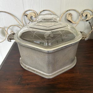 May include: A vintage, square-shaped metal casserole dish with a clear glass lid. The lid features a decorative emblem and a handle. The dish has side handles and a textured, silver-colored exterior. The casserole dish is on a dark wooden surface.