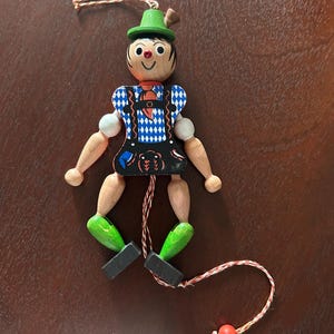May include: A wooden marionette doll with a green hat, blue and white diamond patterned shirt, and black shorts. The doll has wooden arms and legs, with green painted feet. A string is attached for manipulation.