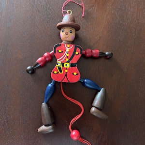 May include: Wooden toy soldier with a red jacket, brown hat, and blue legs. The toy has articulated arms and legs, with a red string for movement. The soldier's uniform features gold accents and a black belt. A classic, handcrafted toy.
