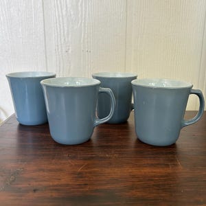 May include: A set of four matching mugs in a solid dusty blue color. Each mug features a smooth, rounded body with a sturdy handle on the side and a white interior. The mugs are arranged in a group, showing their uniform shape and consistent blue finish.