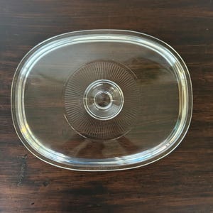 Könnte beinhalten: Ein klarer, ovaler Glasdeckel mit einem zentralen, runden Knaufgriff. Die Mitte des Deckels ist mit einem strahlenförmigen Muster aus feinen, eingravierten Linien verziert, die vom Knauf ausgehen und von einem glatten, transparenten Rand umgeben sind.