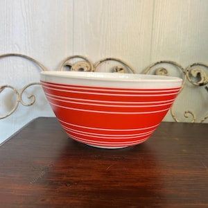 May include: A vintage red and white mixing bowl with horizontal white stripes. The bowl has a rounded shape and a white interior. It is sitting on a dark wooden surface.