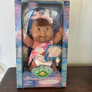 May include: A Cabbage Patch Kids Millennium Celebration doll in its original packaging. The doll has brown hair, a pink bow, and a sash that reads "2000". The packaging features a cityscape with fireworks and the text "Collector Edition".