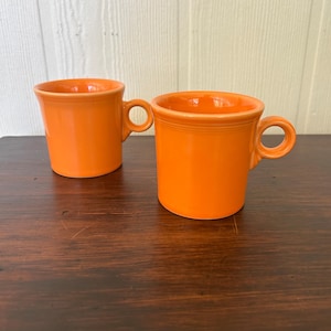 Könnte beinhalten: Zwei leuchtend orangefarbene Keramikbecher mit glatter, glänzender Oberfläche. Jeder Becher hat einen zylindrischen Körper, einen leicht ausgestellten Rand und einen kleinen, kreisförmigen Henkel an der Seite. Die Becher haben eine durchgehend kräftige Farbe.