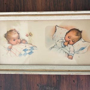May include: A framed vintage print featuring two illustrations of babies. One baby is awake, and the other is sleeping. The frame is rectangular with a distressed white and gold finish. The artwork is in soft pastel colors.