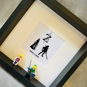 May include: A black framed picture with a white mount and a black and white silhouette of two figures holding swords. The text "SARAH & DAVID" is printed below the figures. Two small figurines are in the foreground, one is green and the other is yellow.