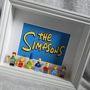 May include: A white framed artwork displaying the words "The Simpsons" in yellow on a blue backdrop. Below the text are Lego figures of the Simpson family and friends. The frame is white.