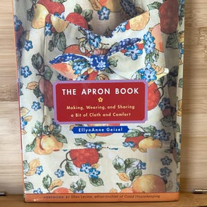 May include: A book titled "The Apron Book" with a cover featuring a floral apron design. The cover text reads "Making, Wearing, and Sharing a Bit of Cloth and Comfort" by EllynAnne Geisel.