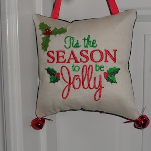 May include: A square, off-white Christmas ornament with the embroidered phrase "Tis the SEASON to be Jolly" in red and green. The ornament has holly accents, a red ribbon for hanging, and two red jingle bells.