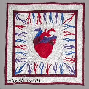 Puede incluir: Una colcha blanca con un corazón rojo, azul y morado en el centro. El corazón está rodeado de ramas rojas, azules y moradas. La colcha está bordeada con un ribete marrón. El texto "@ByNessie505" está en la esquina inferior derecha.