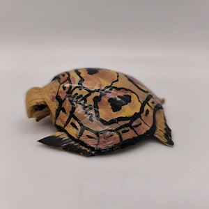 May include: A small turtle figurine crafted from natural fibers, featuring a painted shell with a tan and yellow base color, accented by distinct black outlines and markings that define the scutes across the curved surface.