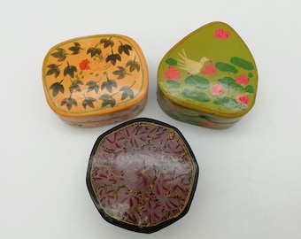 Könnte beinhalten: Drei kleine, kunstvoll bemalte Holzkisten. Eine Kiste ist gelb mit grünen Blättern und einer roten Blume, eine andere ist grün mit rosa Blüten und einem weißen Vogel, und die dritte ist schwarz mit violetten und goldenen Blumendesigns.