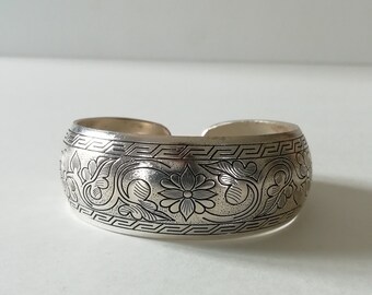 May include: A silver cuff bracelet with an intricate floral design. The design features a repeating pattern of flowers and leaves.