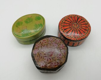 Könnte beinhalten: Drei kleine dekorative Boxen mit bemalten Designs. Eine Box ist oval und grün mit einem floralen Muster. Eine weitere Box ist rund und schwarz mit einem roten und orangefarbenen floralen Muster. Die dritte Box ist achteckig und schwarz mit einem rosa und goldenen floralen Muster.