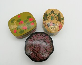 Könnte beinhalten: Drei kleine dekorative Boxen mit bemalten Designs. Eine Box ist grün mit einem weißen Vogel und rosa Blumen, eine ist gelb mit einer Szene von Menschen und einer Kutsche, die von einem Pferd gezogen wird, und eine ist schwarz mit rosa und lila Blumen.