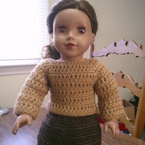 May include: A doll wearing a handmade crochet outfit. The doll has brown hair and is dressed in a tan crochet sweater and a dark brown crochet skirt. The outfit is a simple, classic design.