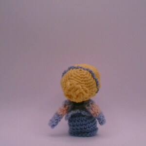 May include: A small, handmade crochet doll seen from behind. The doll features a yellow head with blue trim, a blue body, and arms with pink and blue details. The doll is set against a plain background.