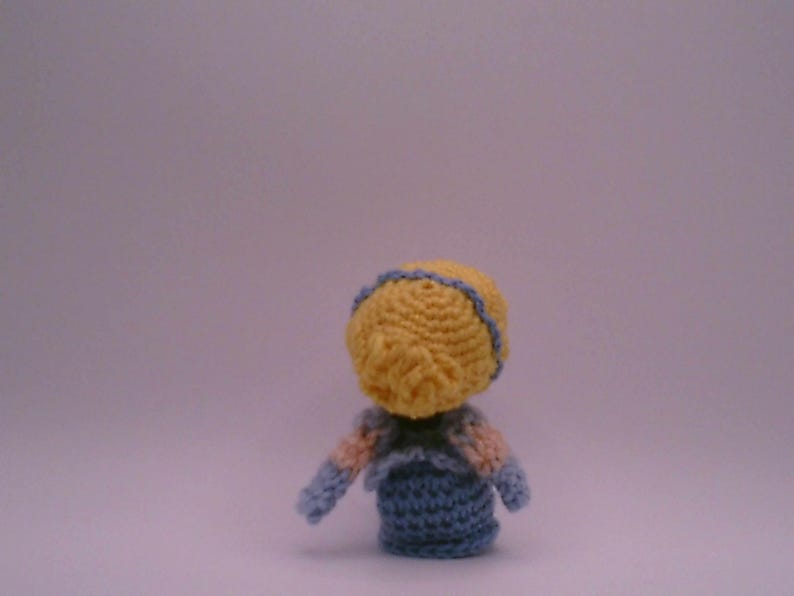 May include: A small, handmade crochet doll seen from behind. The doll features a yellow head with blue trim, a blue body, and arms with pink and blue details. The doll is set against a plain background.