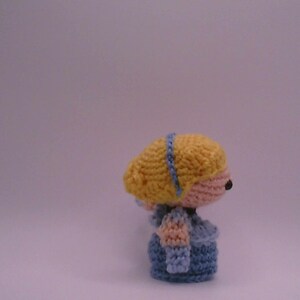 May include: Handmade crochet doll featuring a yellow head with a blue headband, a pink face, and a blue outfit. The doll has a small black nose and a blue dress with sleeves. A handcrafted item.