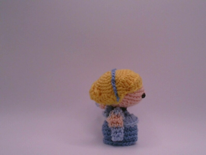 May include: Handmade crochet doll featuring a yellow head with a blue headband, a pink face, and a blue outfit. The doll has a small black nose and a blue dress with sleeves. A handcrafted item.