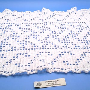 May include: A white, rectangular, crocheted doily with a detailed openwork pattern. The edges are scalloped. A small tag at the bottom reads "Made Exclusively for you by Sharon's Chateau".