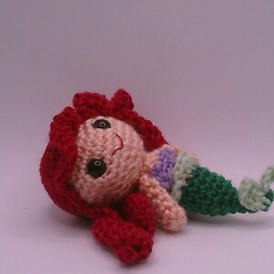 May include: A handmade crochet mermaid doll with red hair, a peach face, and a green tail. The doll has black eyes and a small smile. The mermaid is lying on its side, with its tail curled up.