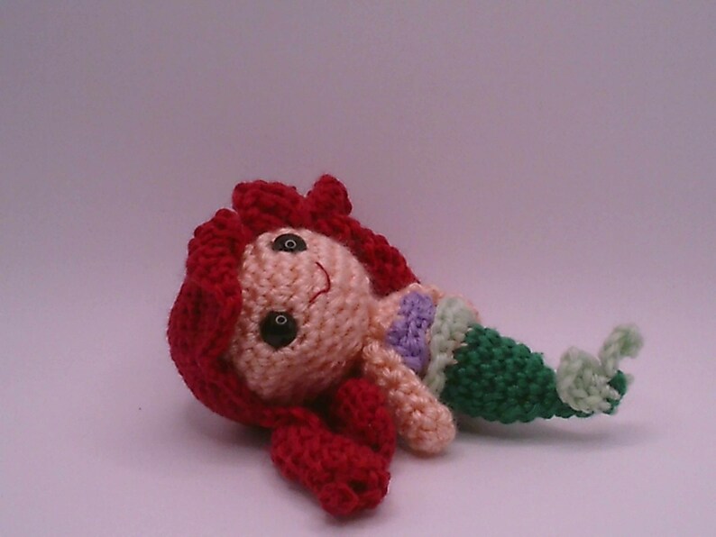 May include: A handmade crochet mermaid doll with red hair, a peach face, and a green tail. The doll has black eyes and a small smile. The mermaid is lying on its side, with its tail curled up.