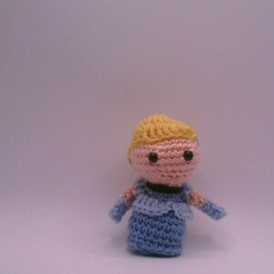 May include: A handmade crochet doll with a light pink face, black eyes, and yellow hair. The doll is wearing a blue dress with matching sleeves. The doll is standing against a plain background.