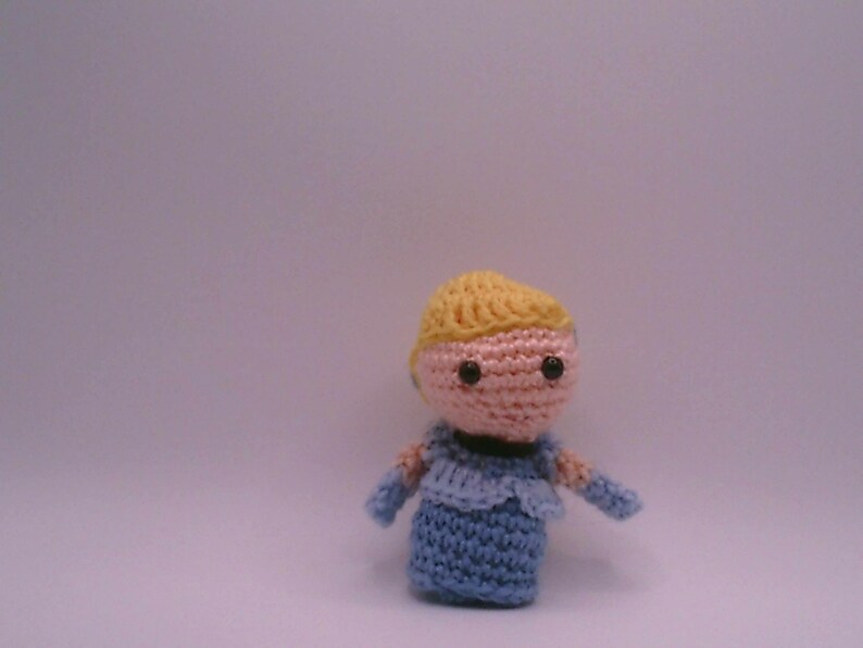 May include: A handmade crochet doll with a light pink face, black eyes, and yellow hair. The doll is wearing a blue dress with matching sleeves. The doll is standing against a plain background.
