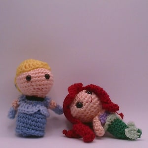 May include: Two crocheted dolls: one in a blue dress with blonde hair, and another as a mermaid with red hair and a green tail. The dolls are handmade and appear to be inspired by fairy tale characters.