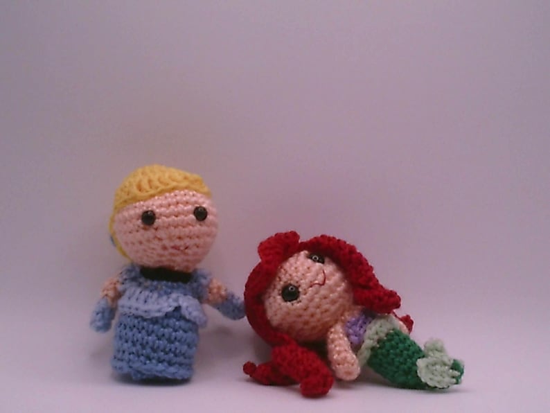 May include: Two crocheted dolls: one in a blue dress with blonde hair, and another as a mermaid with red hair and a green tail. The dolls are handmade and appear to be inspired by fairy tale characters.