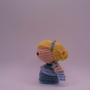 May include: A handmade crochet doll with a light pink face, yellow hair, and a blue headband. The doll is wearing a blue dress and has light pink arms. The doll is a side view.