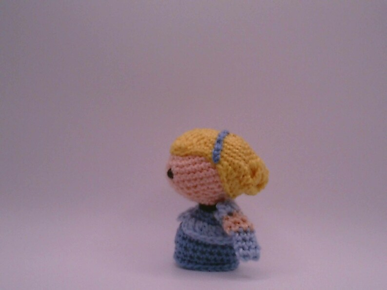 May include: A handmade crochet doll with a light pink face, yellow hair, and a blue headband. The doll is wearing a blue dress and has light pink arms. The doll is a side view.