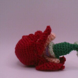 May include: A handmade, crocheted sculpture with a striking red base and a ruffled edge. It features green, cream, and pastel-coloured elements, set against a soft, neutral background. The piece displays a unique, textured aesthetic, showcasing the craft's detail.