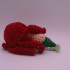 May include: A crocheted octopus in vibrant red, with a cream and green hat. The octopus is lying on its side, with its tentacles curled. The hat is green with a cream-coloured brim. The background is a solid, light purple colour.