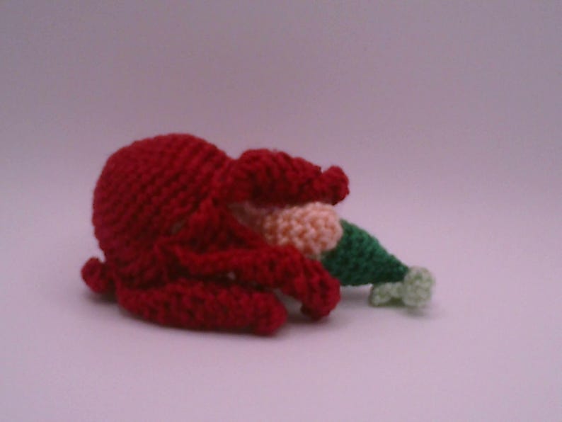 May include: A crocheted octopus in vibrant red, with a cream and green hat. The octopus is lying on its side, with its tentacles curled. The hat is green with a cream-coloured brim. The background is a solid, light purple colour.