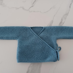May include: A light blue knitted baby cardigan with long sleeves and a wrap-around closure. The cardigan has a tie at the waist.