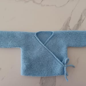 Puede incluir: Un cárdigan de punto azul claro para bebé con estilo envolvente y cierre de nudo.