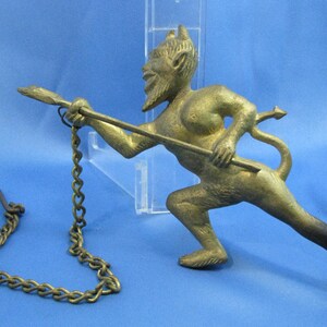 May include: A brass-colored metal figurine of a horned, bearded figure in a crouching pose, holding a long spear-like rod attached to a metal chain with a hook at the end. The figure has a pointed tail and is depicted in a dynamic, forward-leaning stance.