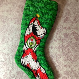 May include: Green Christmas stocking with a Grinch design. The stocking features the Grinch's face and body, with the word "GRINCH" printed repeatedly on the green background. A red loop is attached for hanging.