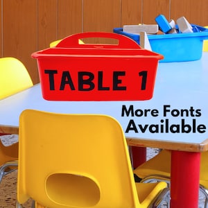 May include: Red plastic bin with a black handle and the text "TABLE 1" in black lettering. The bin is sitting on a light blue table with yellow chairs. The text "More Fonts Available" is written in black on the table.