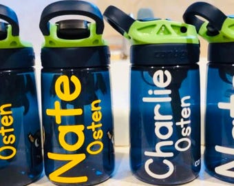 May include: A row of personalized water bottles. The bottles are dark blue with black and green lids. The names "Nate" and "Charlie" are printed in yellow and white, respectively, with the last name "Osten" below each name. The bottles are designed for kids.