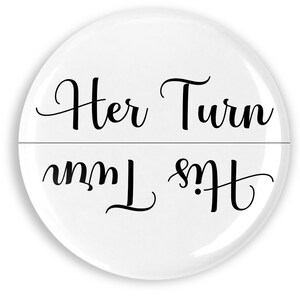 May include: A white, round button with the words "Her Turn" above and "His Turn" below, in black cursive script. The text is divided by a horizontal line. The button has a glossy finish.