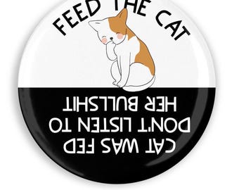May include: A round button with a white and black split design. The top half has the text "FEED THE CAT" and a cartoon cat with brown and white fur. The bottom half reads "CAT WAS FED DON'T LISTEN TO HER BULLSHIT".