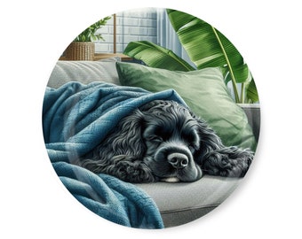May include: A black cocker spaniel dog is sleeping on a grey sofa, covered by a blue blanket. The dog is curled up on a cushion with a green cover. The sofa is in a room with a window and a plant in the background.